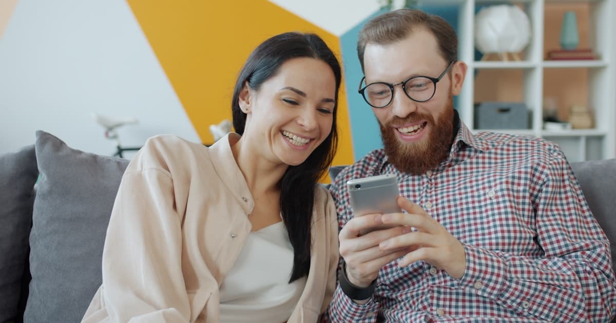 Couple laughing while looking at a smartphone together — reviewing their shared spending