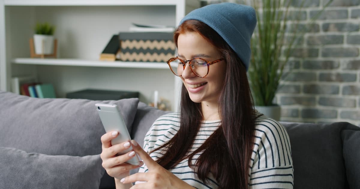 Young woman wearing a beanie and glasses scrolling on her phone while relaxing on a sofa