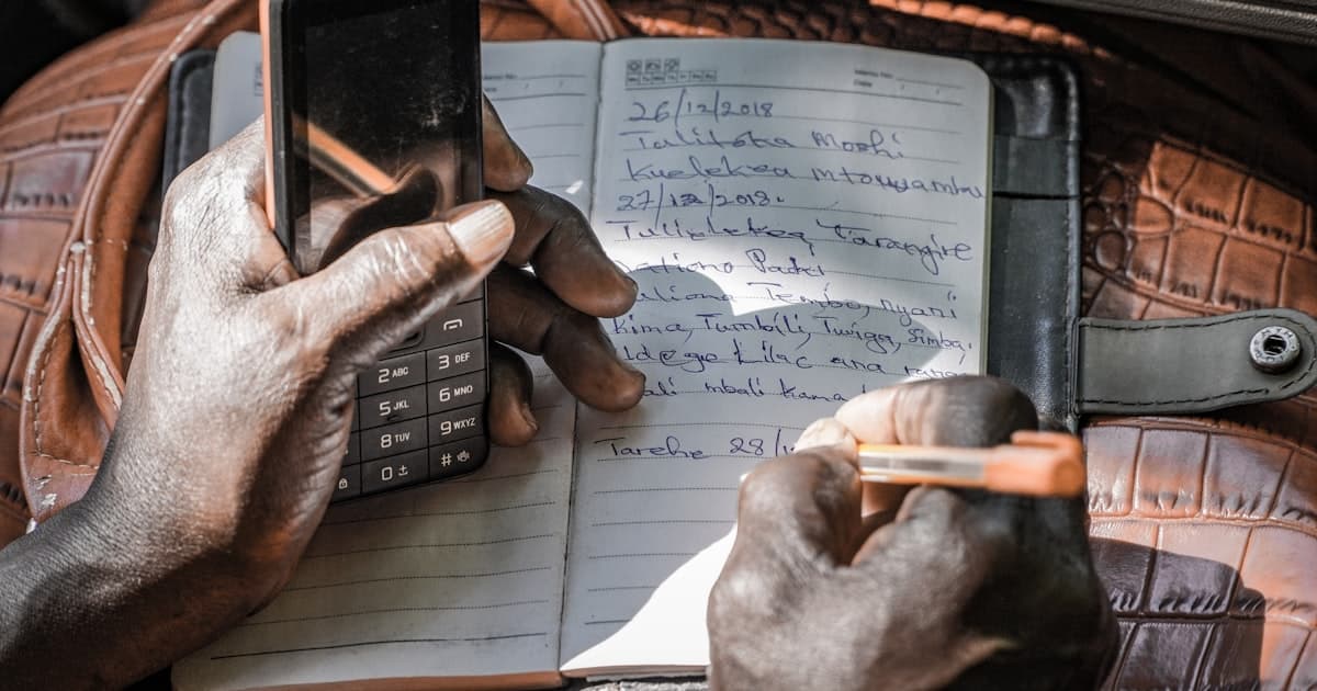 A person writing notes in an open notebook while holding a phone