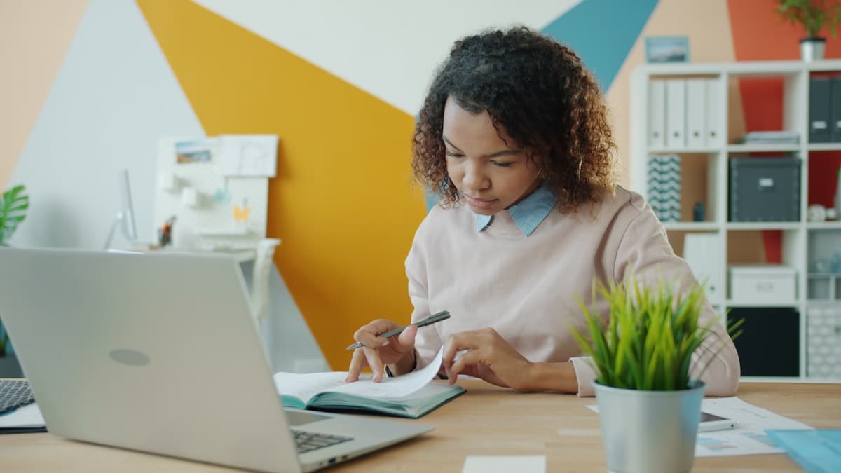 A woman working at a wooden desk with a laptop and open notebook, reviewing her finances