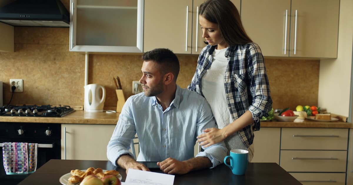 man reading bills at kitchen table while his wife stands beside him supportively